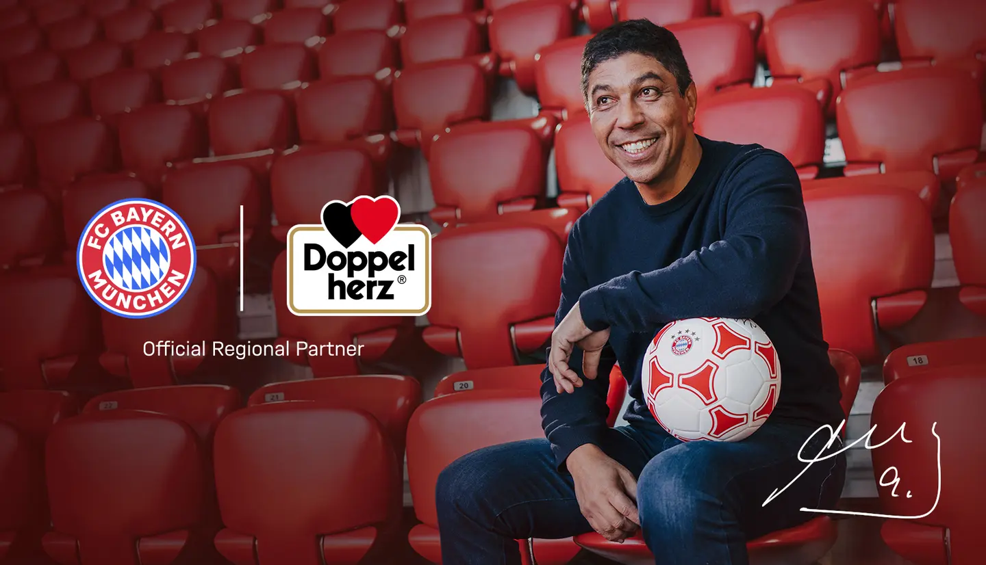A smiling man sits in a stadium, holding a soccer ball. The background features red seats, and logos for FC Bayern München and Doppelherz are displayed, indicating sponsorship. The image conveys a partnership in a sports context.