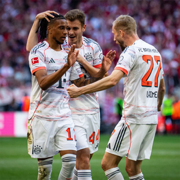 Three soccer players from Bayern Munich celebrate a successful play. One player, wearing jersey number 14, gestures enthusiastically while being embraced by teammates. The background shows a cheering crowd, adding to the festive atmosphere of the moment.