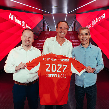 Three men stand in a corridor at Allianz Arena, smiling and holding up a red football jersey. The jersey displays "FC Bayern Munchen," "2027," and "DOPPELHERZ" on the back. The setting features red lighting and decorative steps leading deeper into the venue.