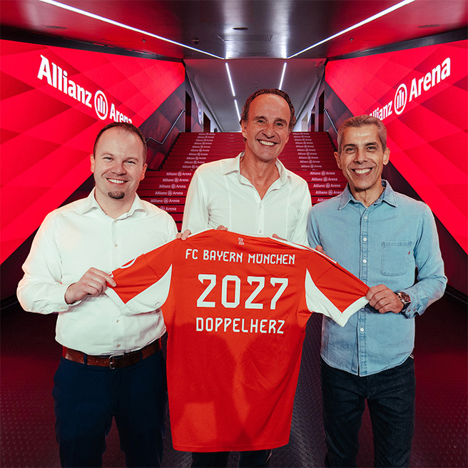 Three men stand in a corridor at Allianz Arena, smiling and holding up a red football jersey. The jersey displays "FC Bayern Munchen," "2027," and "DOPPELHERZ" on the back. The setting features red lighting and decorative steps leading deeper into the venue.