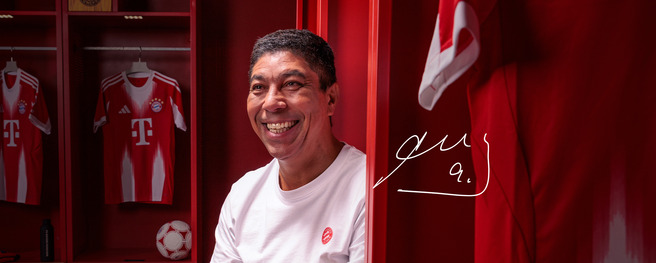 A smiling man in a white t-shirt sits in a red locker room with Bayern Munich football jerseys hanging behind him. A football is on the floor, and a signature appears beside him. The overall atmosphere is inviting and enthusiastic.