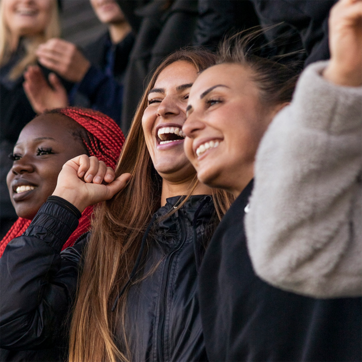 A group of three women is joyfully smiling and cheering. They are dressed in casual, sporty clothing, conveying excitement and happiness. The atmosphere suggests a lively event, with each person actively participating and enjoying the moment together.