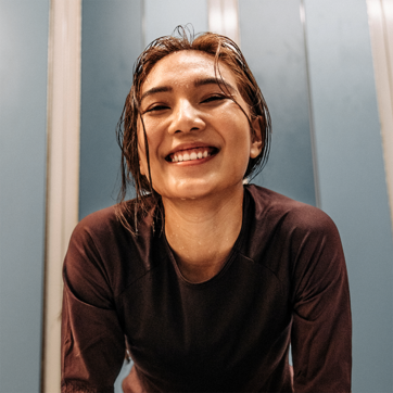 A young woman with wet hair smiles brightly at the camera, exuding a sense of joy and energy. She is wearing a dark athletic shirt, and the background features soft blue tones, contributing to an uplifting atmosphere.