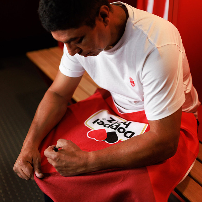 A man in a white shirt sits on a bench in a locker room, attentively adjusting a red towel with a heart design. The setting has wooden benches and subtle lighting, emphasizing a focused, sports-oriented atmosphere.