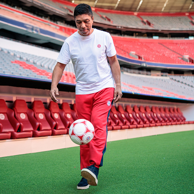 A man in a white t-shirt and red athletic pants is skillfully juggling a soccer ball in a stadium. The empty stands and field are visible in the background. The atmosphere suggests a training or casual practice session.